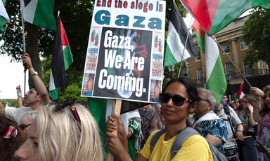 Woman wearing sunglasses holds picket sign reading: "End the siege in Gaza. Gaza, we're coming."