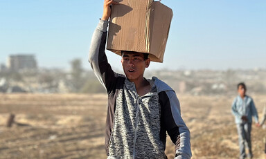 A man in a gray and black top carries a cardboard box on his head.