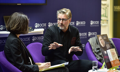 A man wearing dark clothes does a hand gesture as he sits beside another person on a stage 