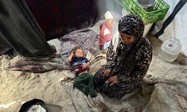 A woman sits on sandy ground next to two small children in a sparsely furnished tent