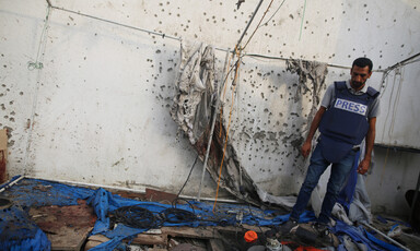 A man in a press vest looks down at the damage after an airstrike. There are shrapnel holes in the wall beside him.