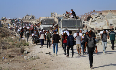 People walk alongside trucks on a dirt road in the daytime, carrying aid. 