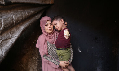 A woman wearing pink prayer clothes holds a toddler, dressed in a red shirt and green shorts. The toddler is missing his left arm.