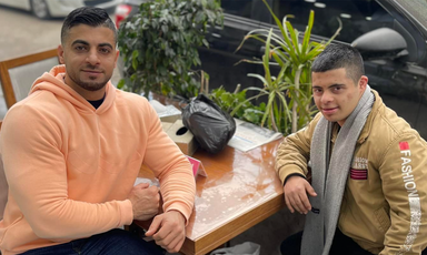 Two men sit outdoors with their elbows resting on a table 