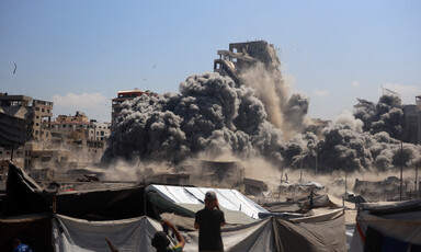 Daytime scene of a building collapsing, giant plumes of gray smoke, with tents and people in the foreground.