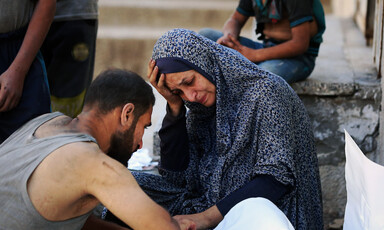 A woman in a blue headscarf holds her head in her hand. A man faces her. They are both looking at a body wrapped in a white shroud.