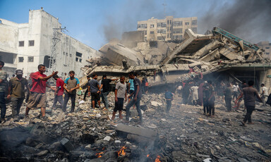 People stand on top of the rubble of a destroyed building, with black smoke rising around them. Several small fires are visible in front of them.