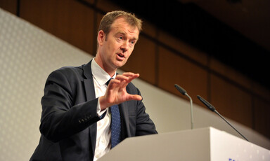 A man wearing a navy suit and white shirt stands at a podium 