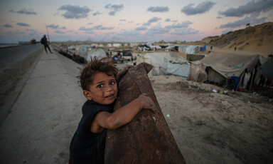 A small child clings to a rusty metal object, looking at the camera. Tent shelters are in the foreground.