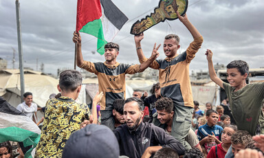 A group of young Palestinian men and children stand in a group. Carried on shoulders, two men hold a Palestinian flag and a symbolic key. Everyone is smiling.