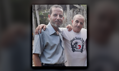 In daytime, two smiling men pose with arms on the other's shoulders.