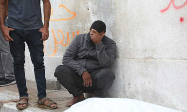 A man in a grey jacket and dark pants crouches against a wall, next to a body wrapped in a white shroud. His hand is covering his face.