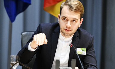 A man wearing a dark jacket and a white shirt sits at a desk with a microphone on it