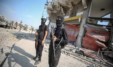 Two armed men stand guard in front of damaged buildings