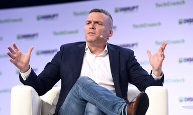 A man with a microphone attached to his face sits on a podium and makes a gesture with his hands 