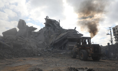 A bulldozer clears rubble from a destroyed house with smoke still billowing in the background