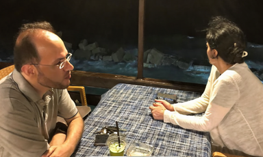 A man and woman lookin different directions while sitting at a table next to the sea at night