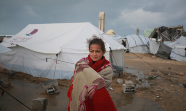 A girl stands in front of a tent shelter. She is dressed in red and wrapped in a blanket