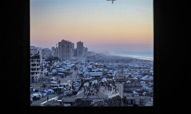 A wide-shot image, showing Gaza's beach crammed with tents with high-rise, damaged buildings in the background during a twilight.