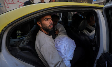 A man sits in the backseat of a yellow car. He is holding a body wrapped in a white shroud.