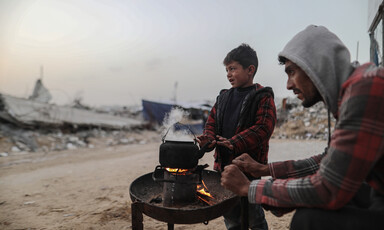 A man and a boy warm their hands over a small tray with a fire and a teapot over it