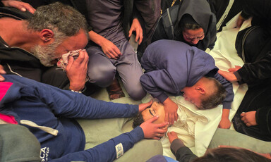 Mourners kneel on the floor, grieving over the body of a youth wrapped in a white shroud