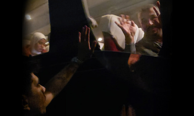 A blurred nighttime shot of a man inside a bus waving and people outside waving.