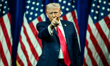 Trump standing in front of row of US flags and points
