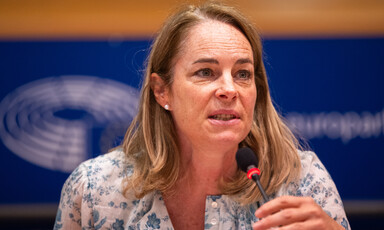 A woman wearing a blouse with a floral pattern speaks into a microphone 