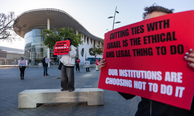 People holding protest signs stand in a plaza outside a modern convention building with curved architecture. 