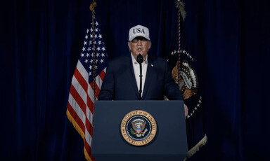 Donald Trump stands at presidential podium speaking and wearing a baseball hat reading "USA"