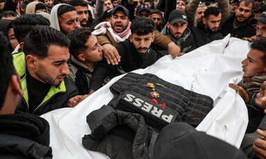 Group of men carries a shrouded body with a bulletproof press vest and flowers on top of it