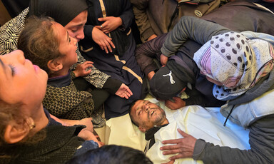 Women and girls mourn over the body of a man wrapped in a white shroud.