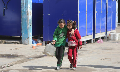 Two girls, one in a green shirt and the other in red, walk together down a street. One is holding a water canister.