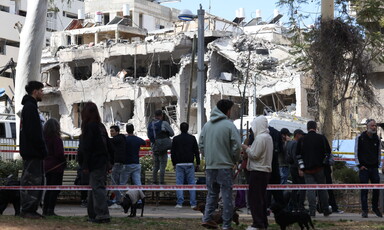 A crowd of people looks up at a partially collapsed apartment building