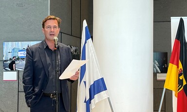 Man holds a piece of paper as he stands behind a microphone stand with flags of Israel and Germany beside him 