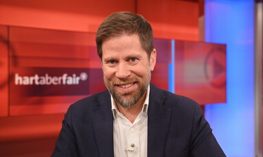 Man wearing a navy jacket and white shirt smiles as he sits in a television studio behind a red backdrop with the words hart aber fair on it. 