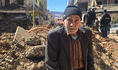 Older man sits in front of rubble of a bombed building