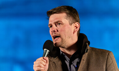 Man speaks into a microphone with a blue backdrop behind him 