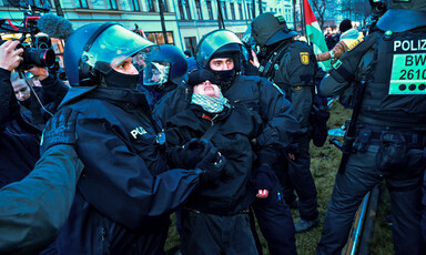 German police in full riot gear grab a protester wearing a checkered scarf by the arms