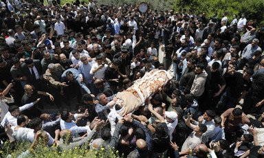 A large crowd of people gather around a body, carried on a stretcher, wrapped in a brown blanket.