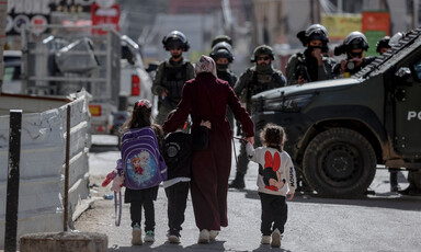 Three children with cartoon backpacks walk on either side of a woman toward armed men in riot gear