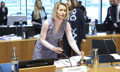 The European Union's Kaja Kallas stands and holds a small bell on a desk bearing papers and refreshments 