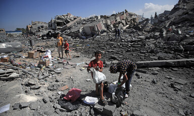 A handful of people inspecting through rubble while a boy, holding a plastic bag, is looking at the camera