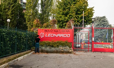 A man stands beside a red sign with the logo of the firm Leonardo and graffiti reading Palestina Libera on it  