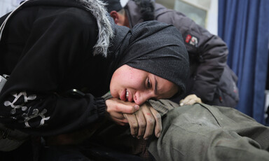 A woman in a black headscarf bends down to place her cheek on the hand of her loved one who was killed