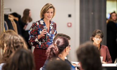 Katharina von Shnurbein, wearing floral print blouse, holds microphone and stands amid a group of sitting people in conference room