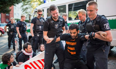 A man wearing a "boycott Israeli apartheid" T-shirt is grabbed by two German police officers. 